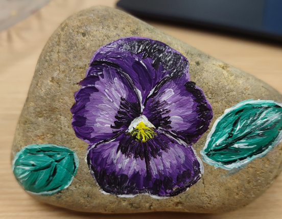 A picture of a rock that has been hand-painted with a purple pansy flower.


